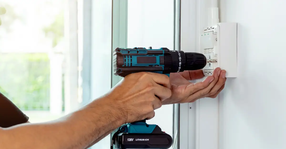 Electrician using a cordless power drill to install or secure a wall-mounted electrical device near a sliding glass door.