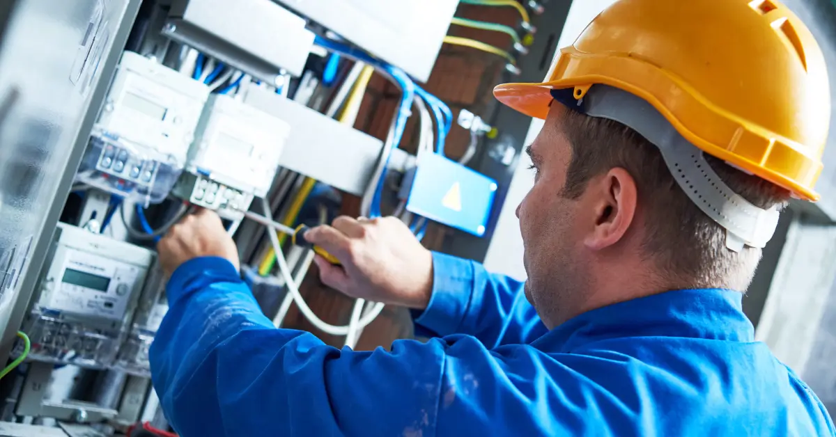 Licensed electrician wearing a hard hat and blue work uniform repairing and rewiring electrical components inside a residential panel.