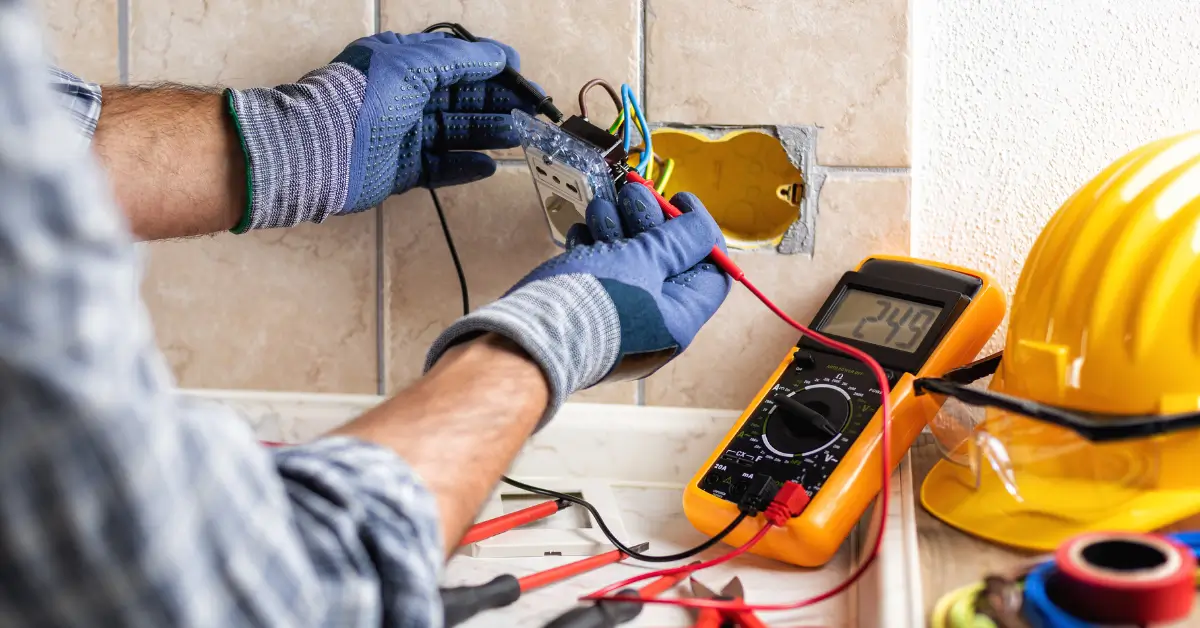 Electrician using a multimeter to test wires while installing or rewiring a wall outlet, with tools and safety gear nearby.
