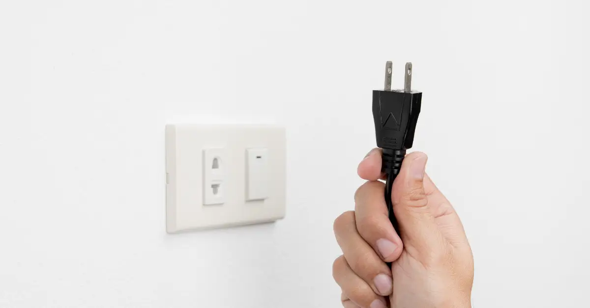 Hand holding a black electrical plug near a wall outlet, illustrating safe plugging and unplugging practices.